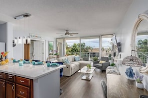 Un amplio salón con un ventanal grande, ventilador de techo y una zona de cocina con un cartel que dice 'BEACH HOUSE'.