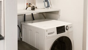 A compact laundry area with a front-loading washing machine, a dryer, and a shelf above containing a first aid kit and other small items.