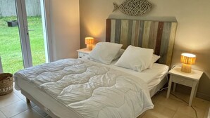 A bedroom with a bed, two bedside tables with lamps, a wall-mounted decorative piece, and a view of the outdoors. - Individual house .  (Le Bois plage)