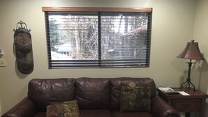 Ein Wohnzimmer mit einem braunen Ledersofa, zwei gemusterten Kissen, einem Holztisch, einem Beistelltisch mit Lampe und einem Fenster mit Jalousien.