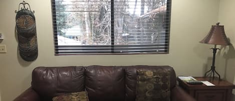 A living room with a brown leather sofa, two patterned cushions, a wooden coffee table, a side table with a lamp, and a window with blinds.