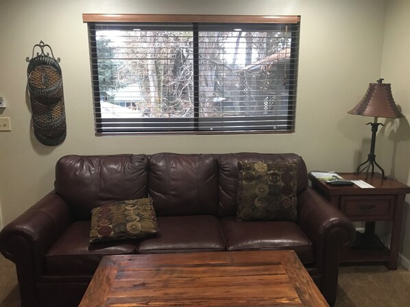 A living room with a brown leather sofa, two patterned cushions, a wooden coffee table, a side table with a lamp, and a window with blinds.