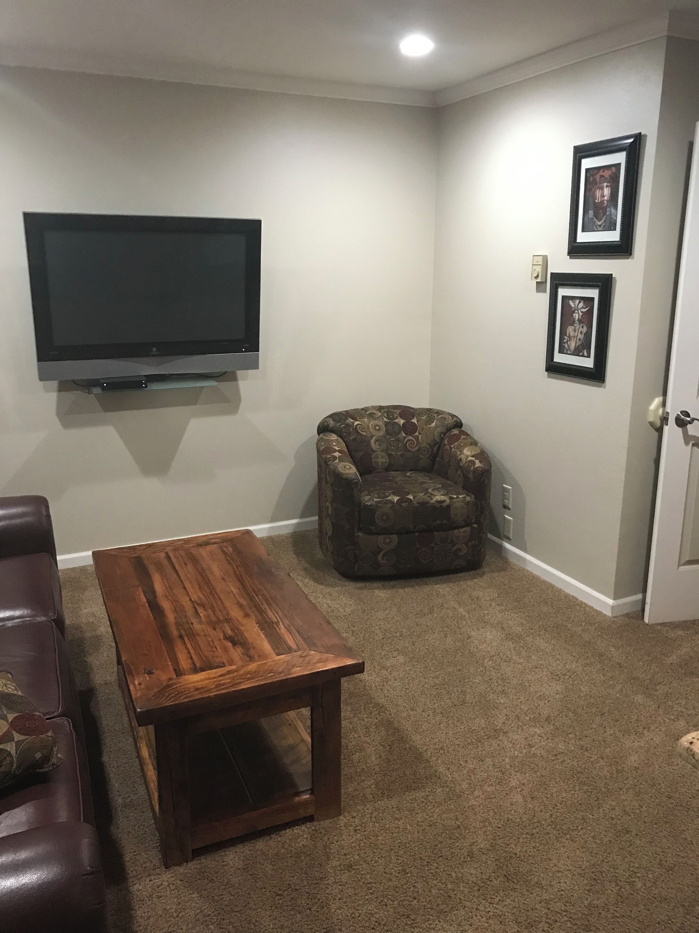 A living room with a flat-screen TV mounted on the wall, a patterned armchair, a wooden coffee table, and a brown carpet.