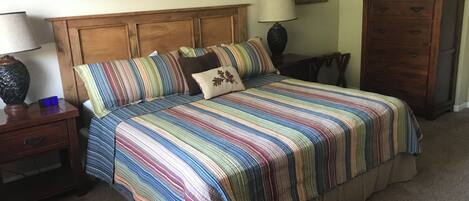 Un dormitorio con cabecera de madera, colcha a rayas, mesita de noche con lámpara y cómoda.