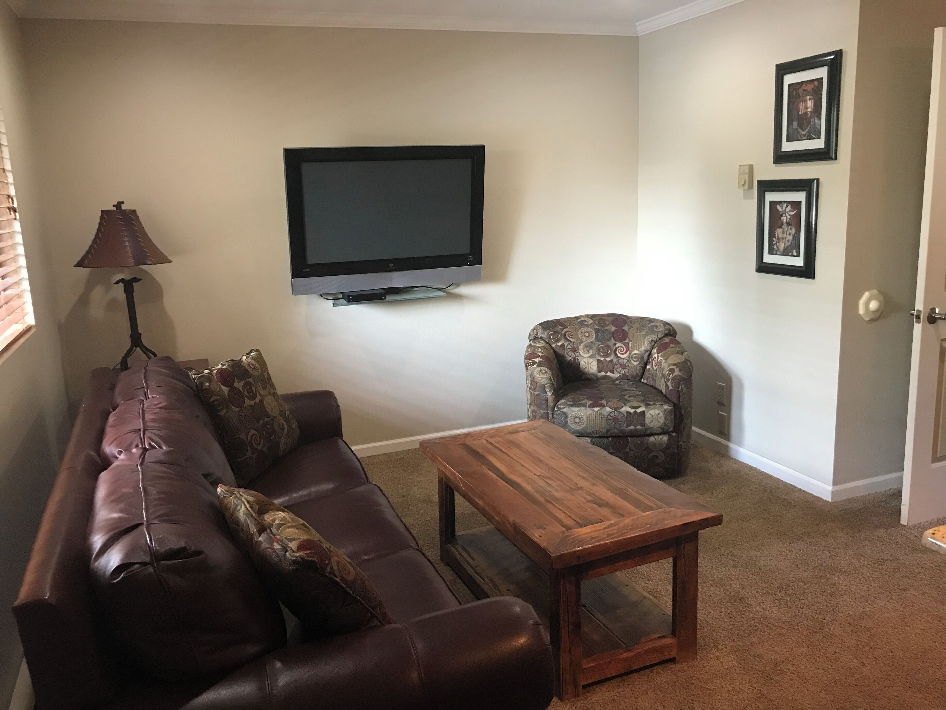 Un salon avec un canapé en cuir, un fauteuil à motifs, une table basse en bois, un téléviseur posé sur un meuble et deux tableaux encadrés accrochés au mur.