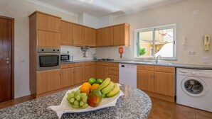 A modern kitchen with wooden cabinets, a built-in oven, microwave, and dishwasher. A fruit bowl with grapes, mango, and bananas is on the granite countertop. - QuietPrivateVilla - superwiffi (Lagos)