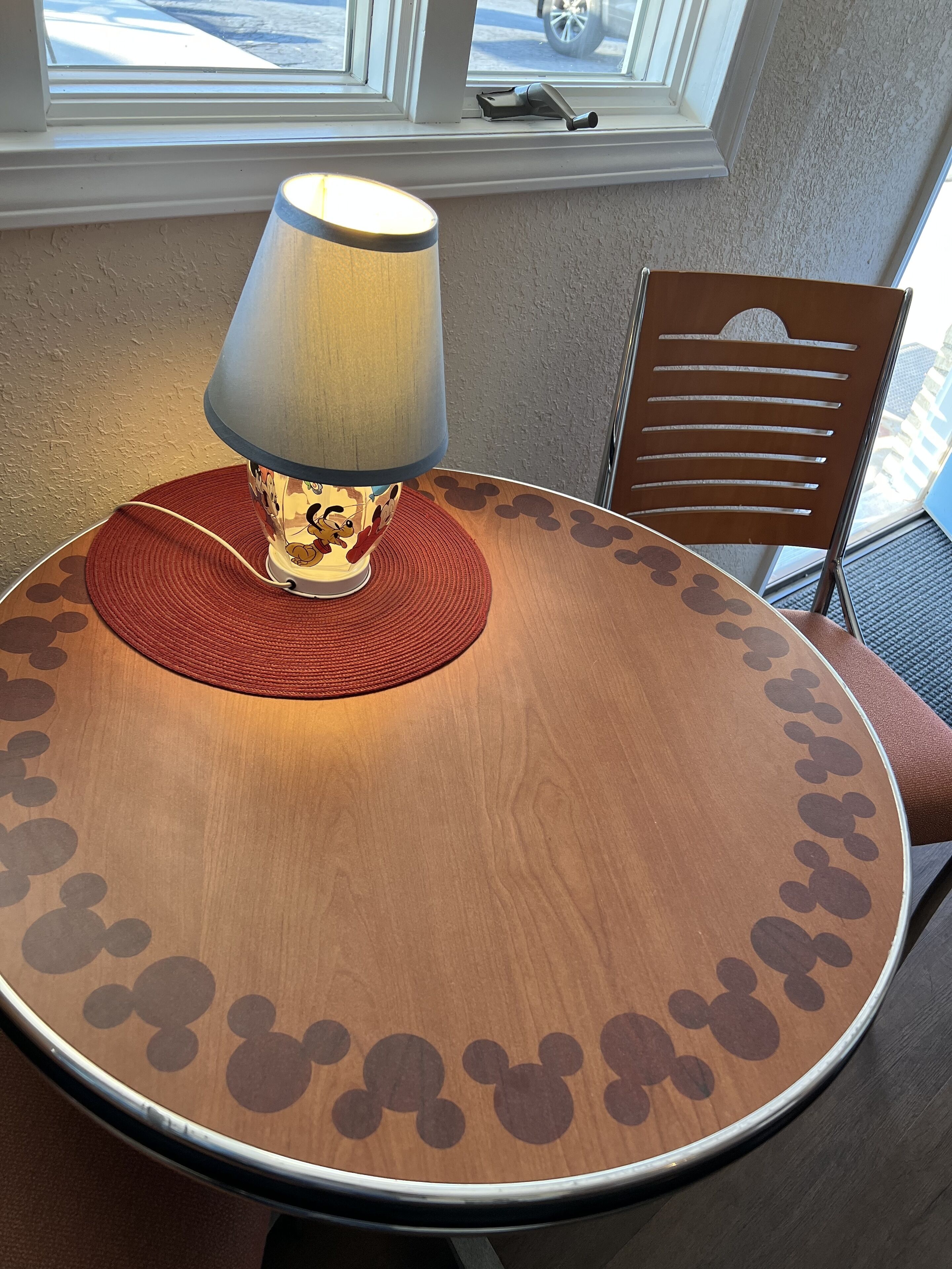 A round table with a Mickey Mouse themed tablecloth, a lamp, and a chair with a Mickey Mouse cutout design.