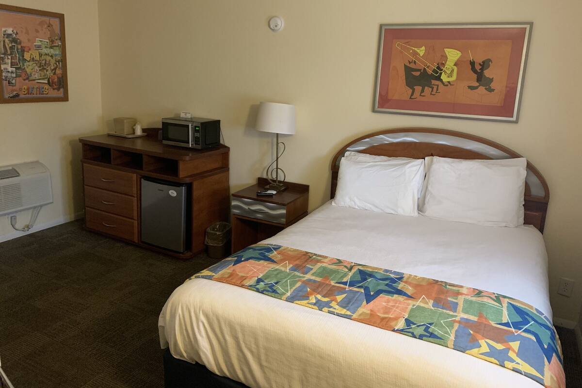 A hotel room with a bed, a desk with a microwave, a lamp, and a trash bin.