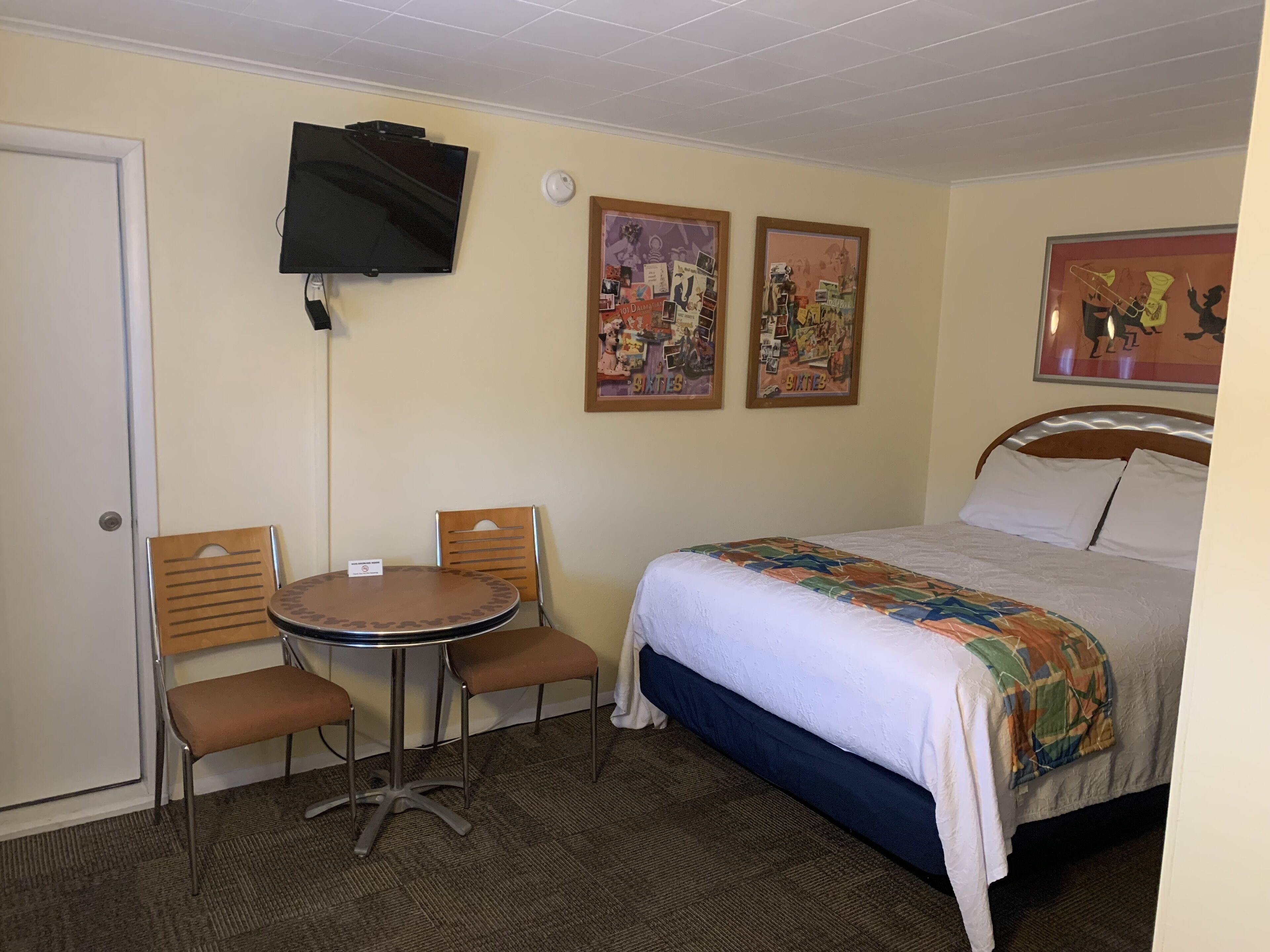 A hotel room with a bed, a TV mounted on the wall, two chairs around a small table, and two framed pictures on the wall.