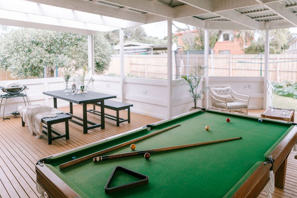 Una mesa de billar con superficie de fieltro verde, rodeada por una terraza de madera y un patio cubierto con una mesa y sillas.