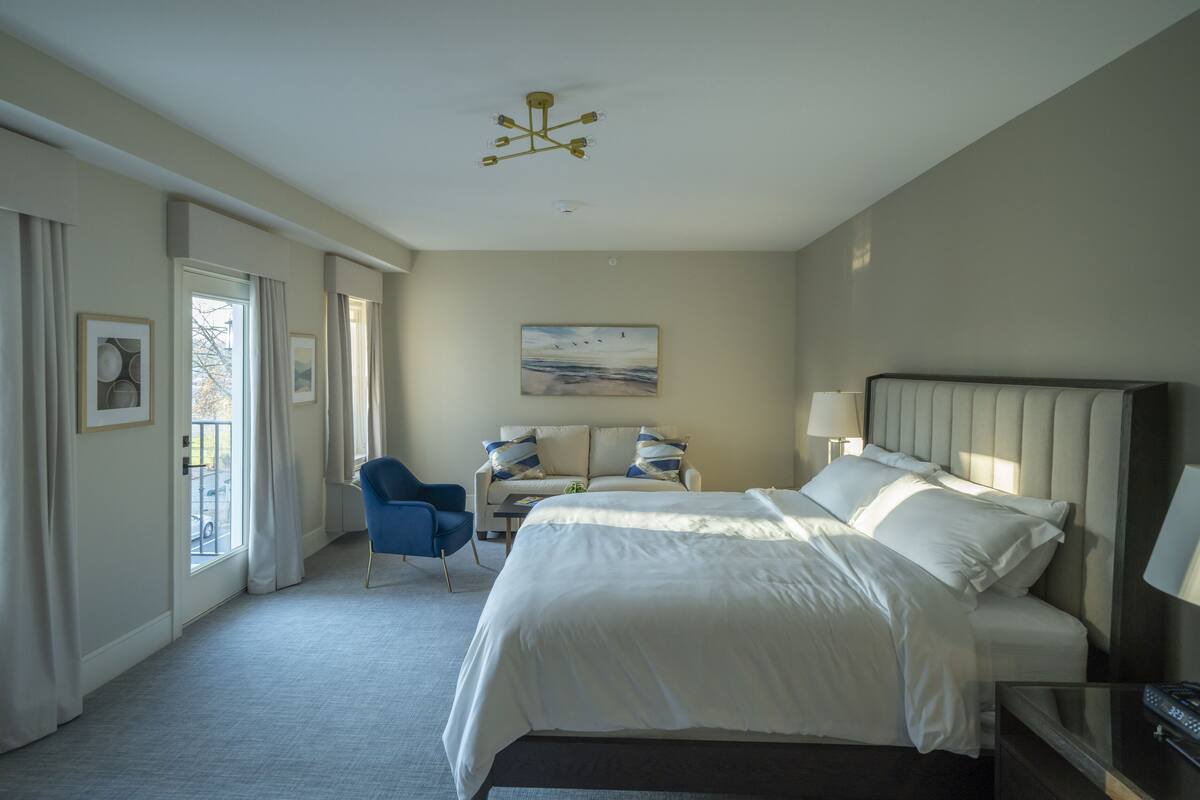 A hotel room with a large bed, a desk with a chair, a window with curtains, and a painting on the wall.