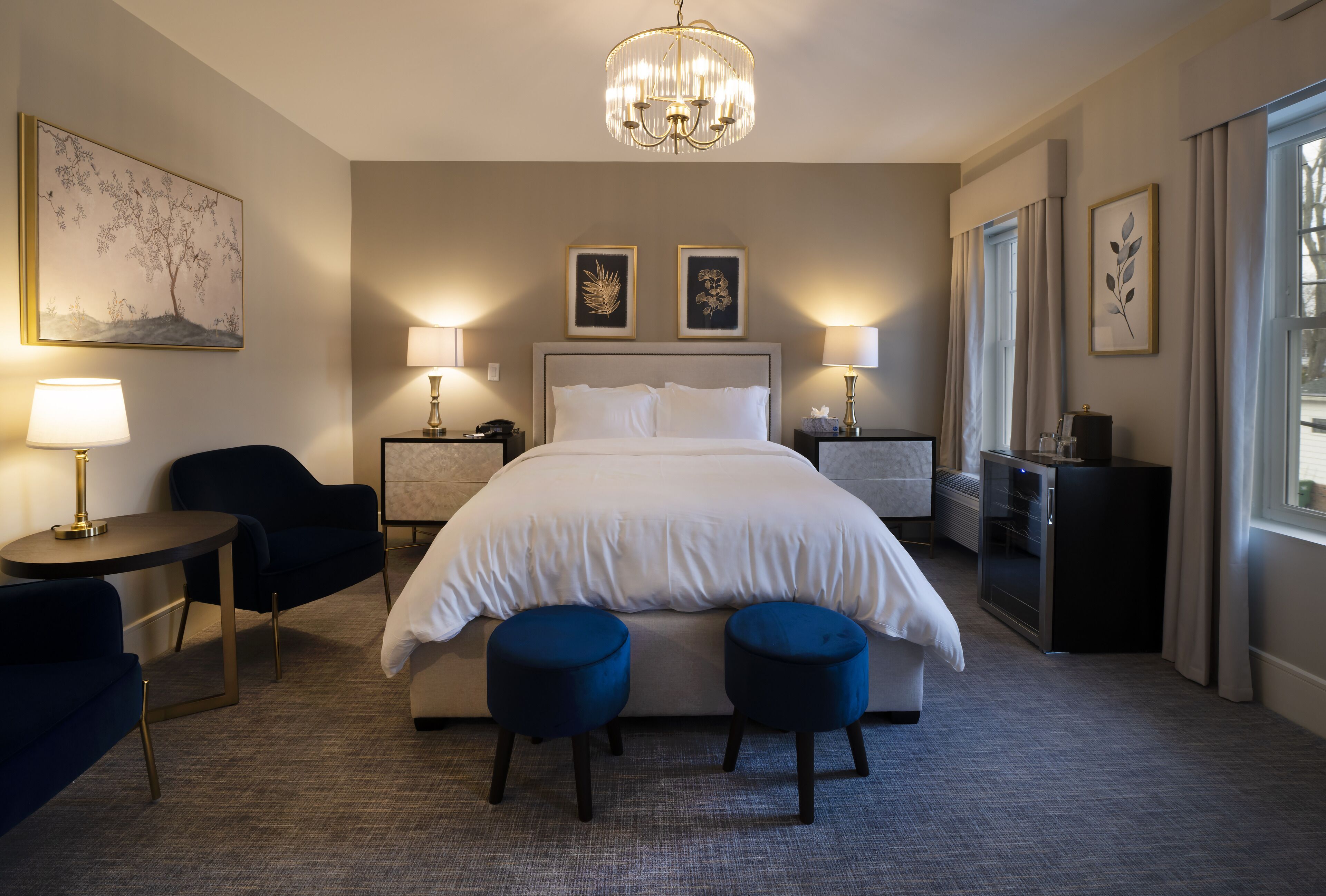 Una camera d'albergo con un letto grande, due comodini con lampade, un tavolino rotondo, una poltrona blu scuro, due poggiapiedi blu, un lampadario e opere d'arte alle pareti.