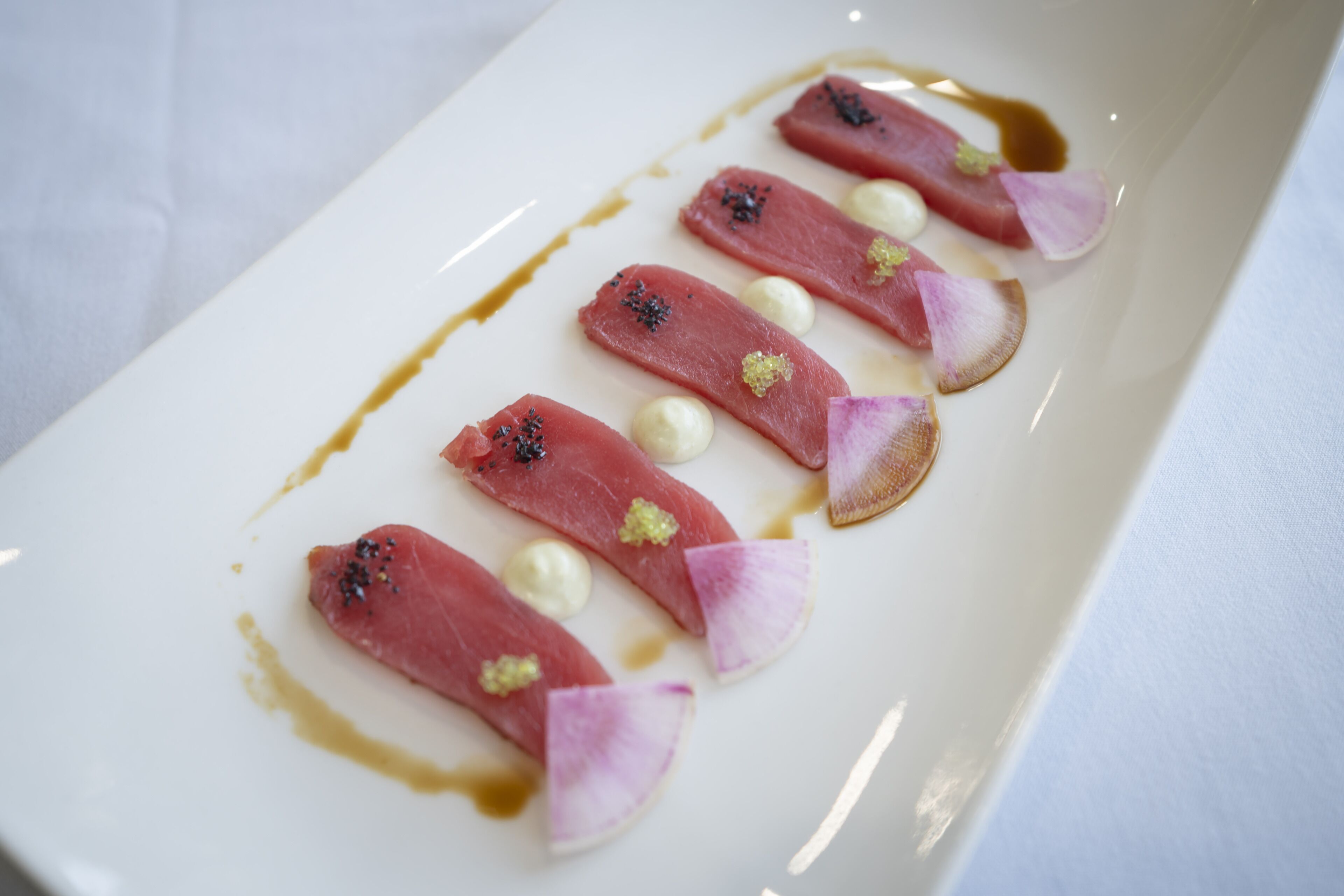 A white rectangular plate with thinly sliced pink meat, white spheres, and pink flower petals, garnished with black and yellow specks.