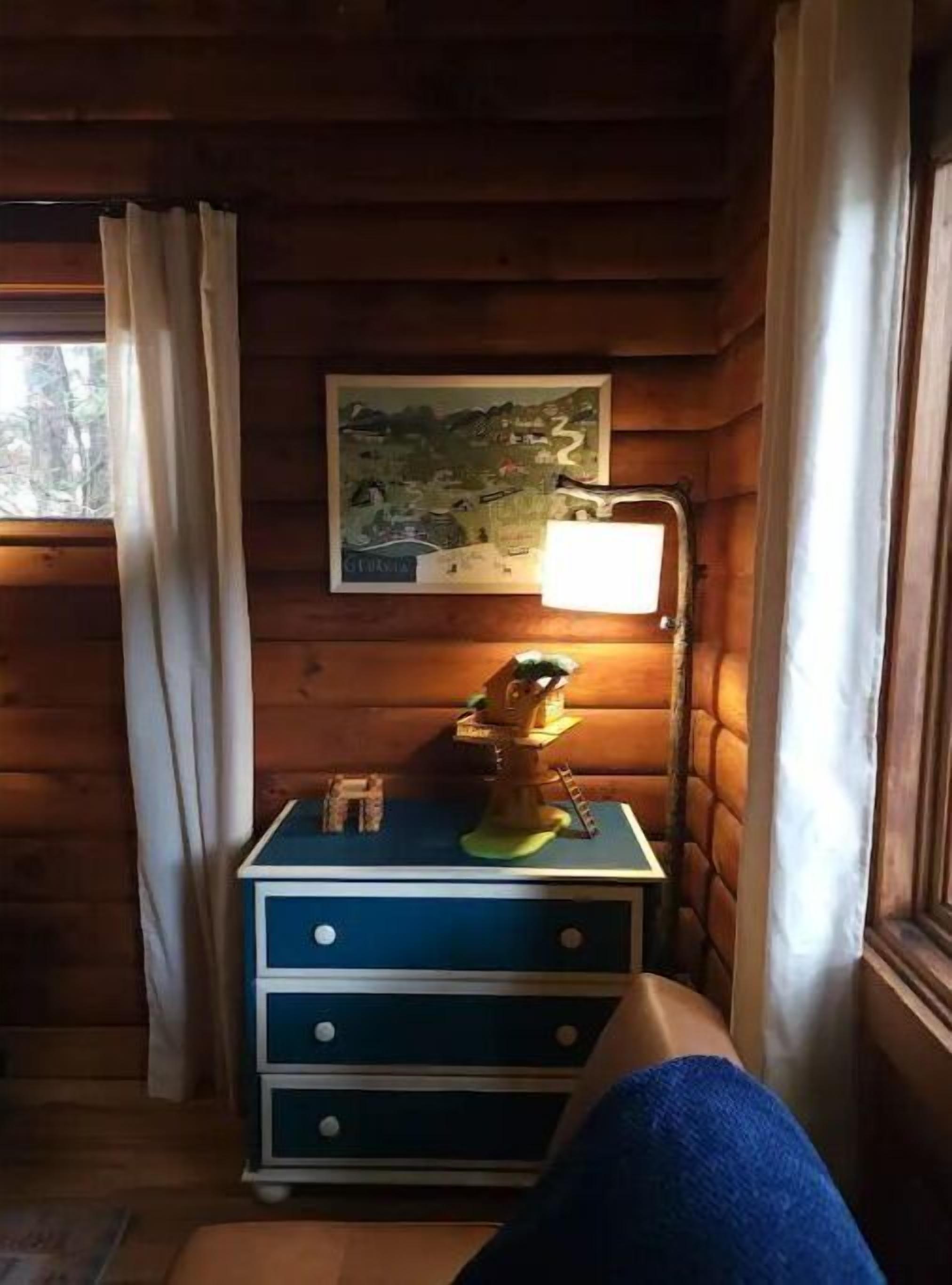 Et hyggeligt sommerhusværelse med seng, kommode, lampe og et kort på væggen.