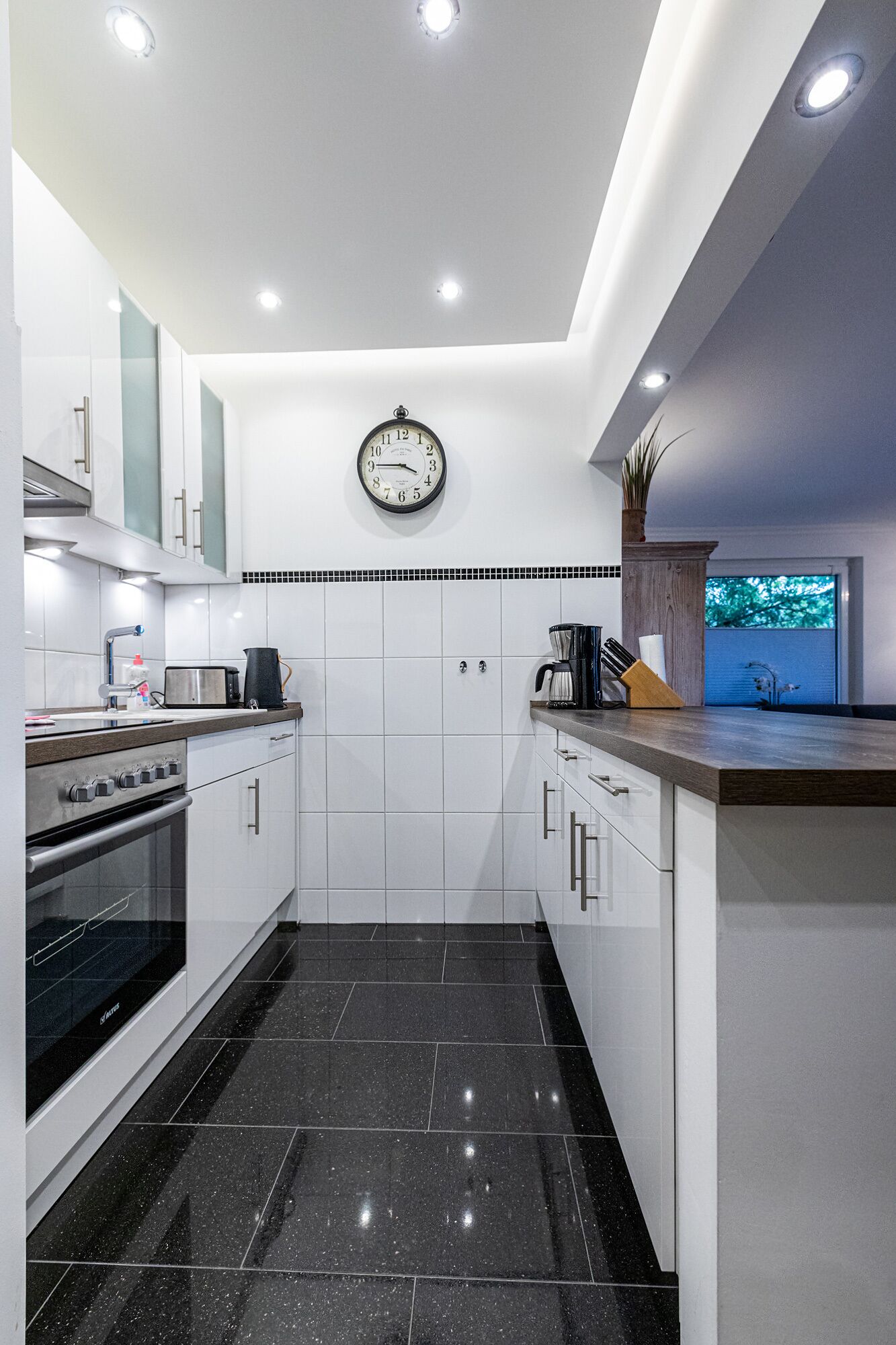 Una cocina moderna con armarios blancos, piso de baldosas negras, un reloj en la pared y una cafetera sobre la encimera.