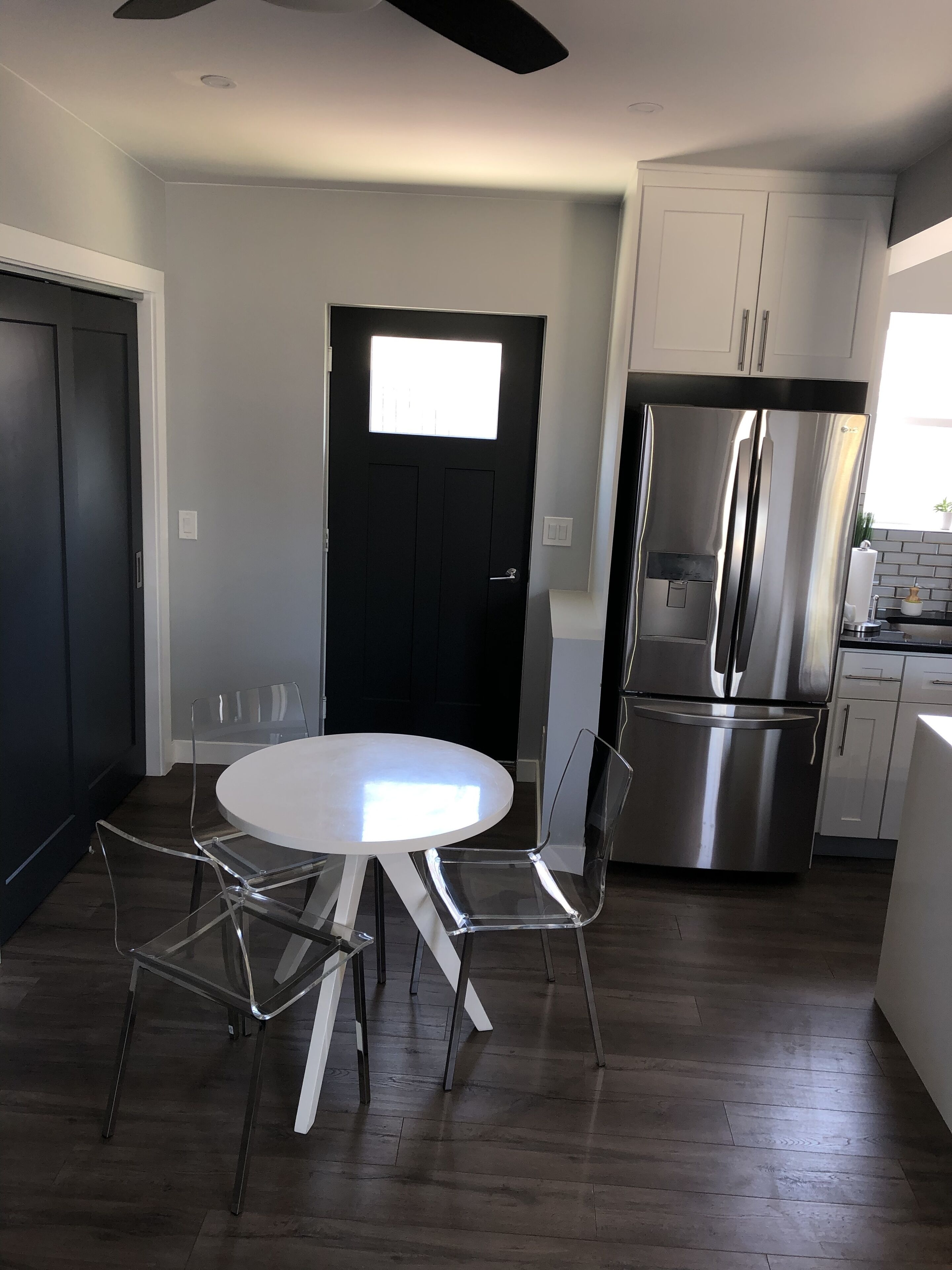 A modern kitchen with a stainless steel refrigerator, a round dining table with clear chairs, and a black door with a window.
