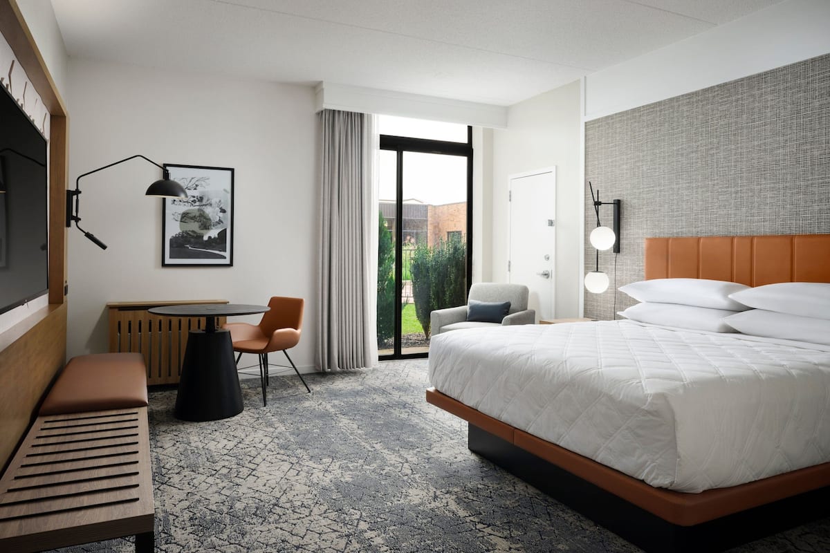 Une chambre d’hôtel avec un grand lit, un bureau avec une chaise, un banc, une lampe fixée au mur et une vue sur la verdure par la porte.