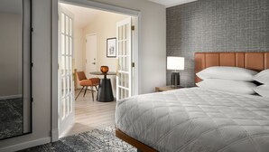 A hotel room with a large bed, a bedside table with a lamp, a chair, and a view of a dining area through an open door. - Sheraton Grand Rapids Airport Hotel (Grand Rapids)