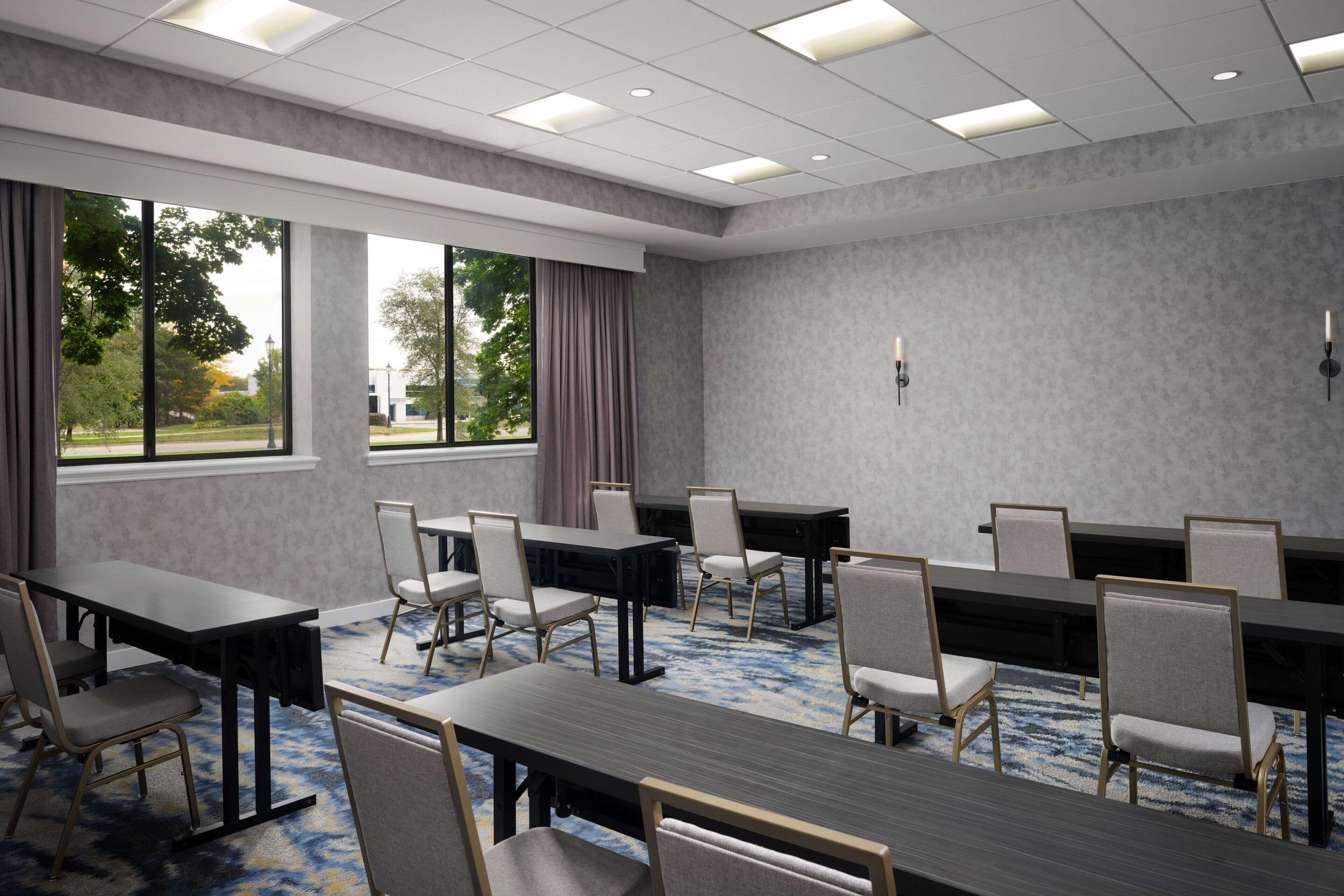 A conference room with multiple tables and chairs, large windows with curtains, and a carpeted floor.