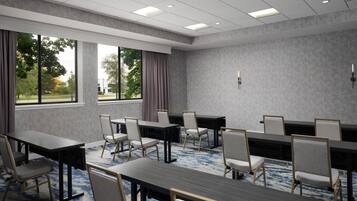A conference room with multiple tables and chairs, large windows with curtains, and a carpeted floor.