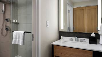 Un baño moderno con cabina de ducha de vidrio, un lavamanos blanco sobre un mueble de madera y un espejo grande que refleja una puerta de madera.