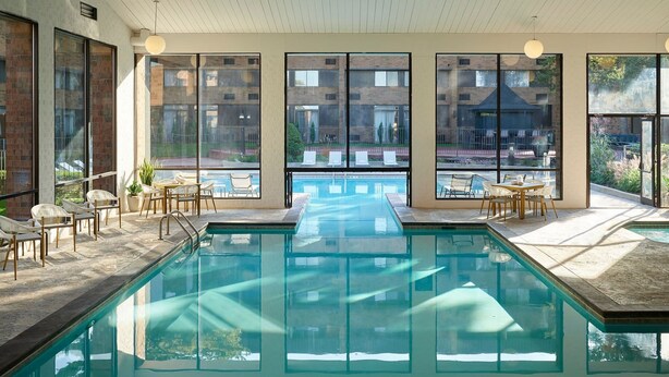 A swimming pool with clear water, surrounded by lounge chairs and tables, large windows offering views of a building and greenery. - Sheraton Grand Rapids Airport Hotel (Grand Rapids)