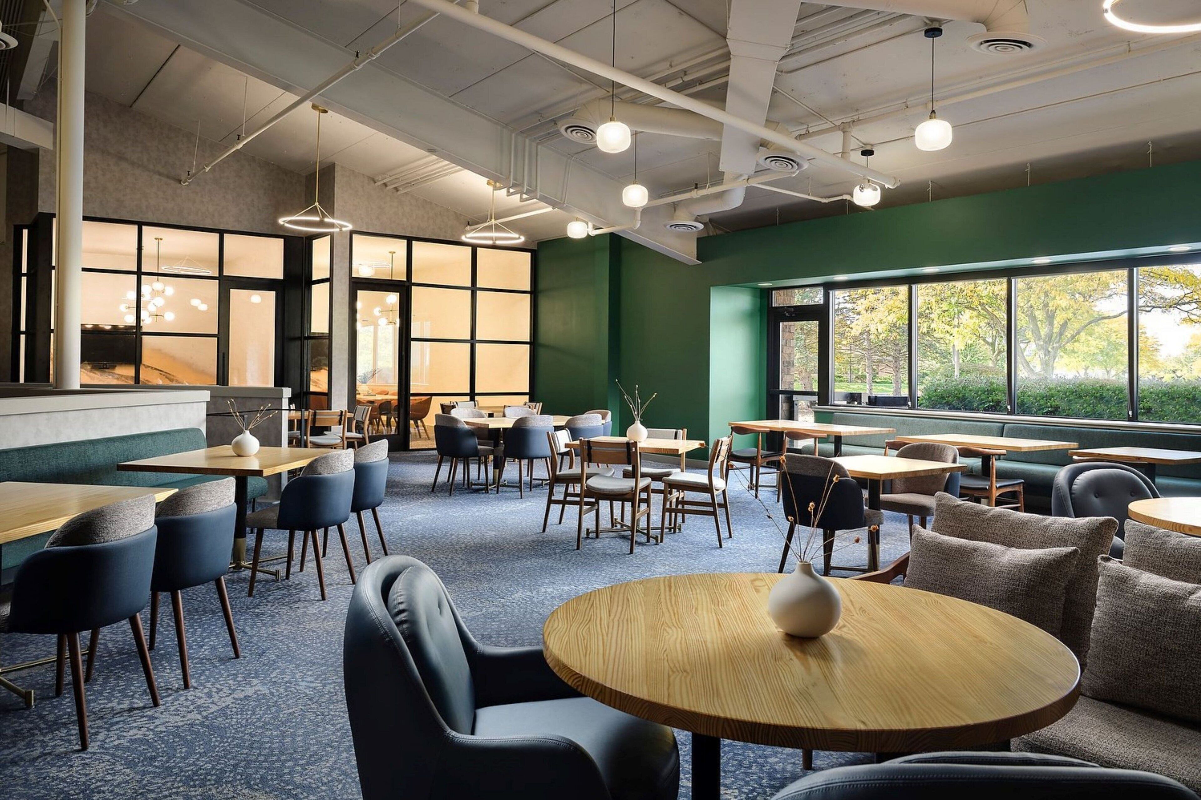 Un espace de restauration moderne avec des tables rondes et des chaises bleues, de grandes fenêtres donnant sur la verdure et un mur d’accent de couleur verte.