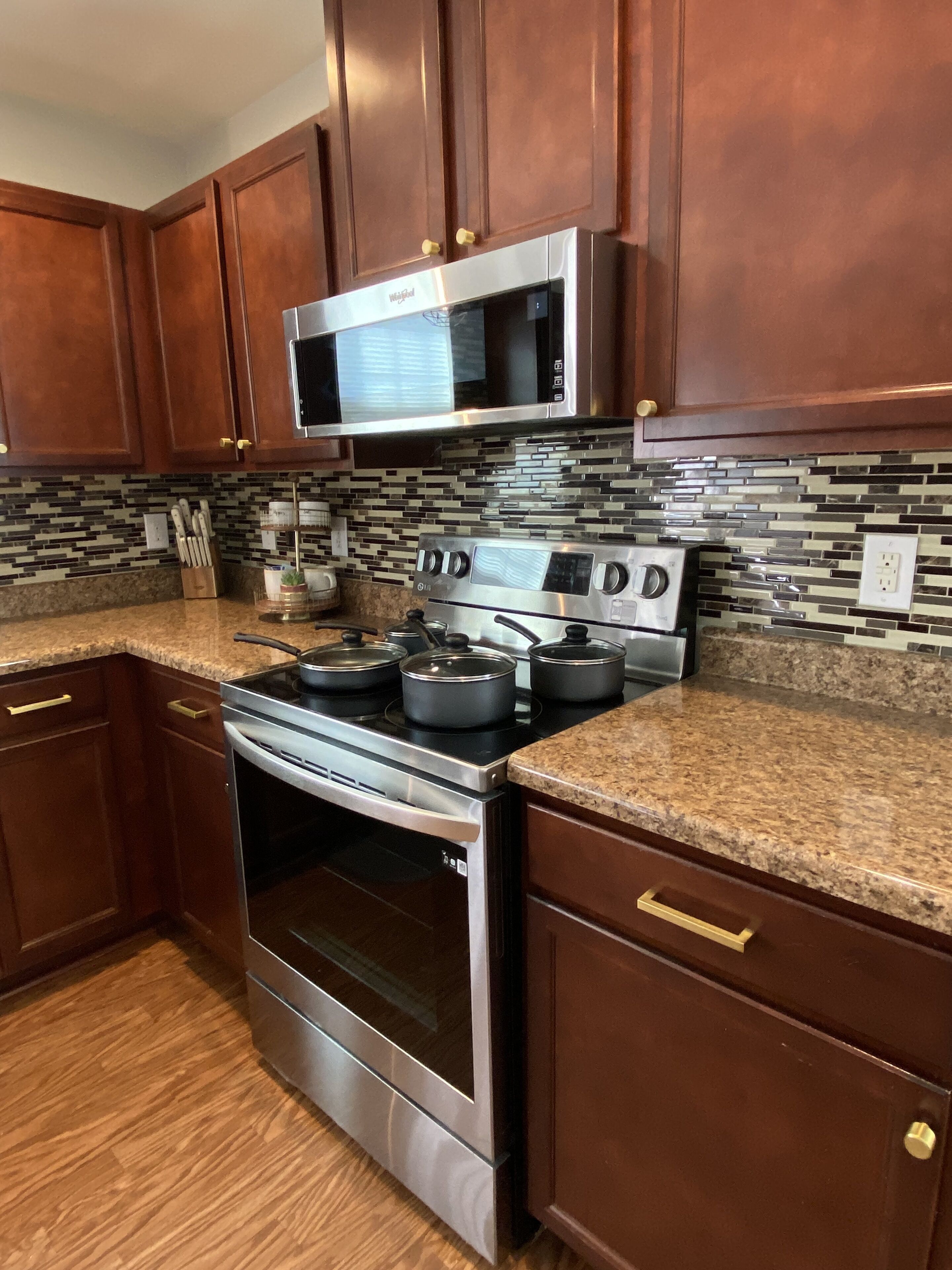 A kitchen with wooden cabinets, a stainless steel microwave, a stove with pots, and a tiled backsplash.