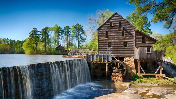 A rustic wooden mill with a water wheel, a stone staircase, and a wooden deck overlooking a calm river. - Beautiful Modern House 5 minutes To Downtown Raleigh (Raleigh)
