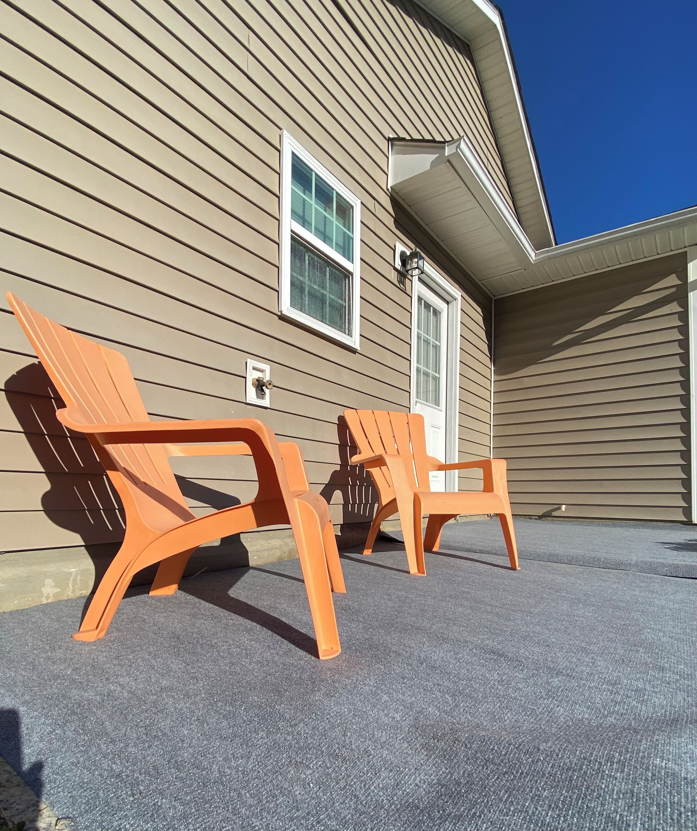 Two orange Adirondack chairs outside a beige house with a window and a door.