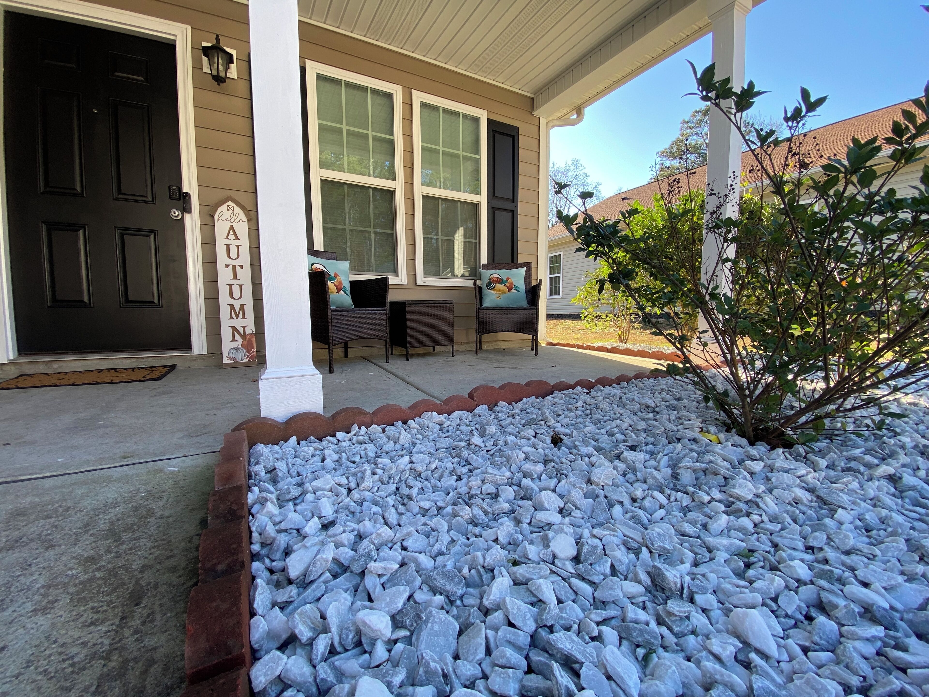 A house entrance with a black door, a welcome sign, a covered porch with wicker furniture, and a garden bed with white pebbles.