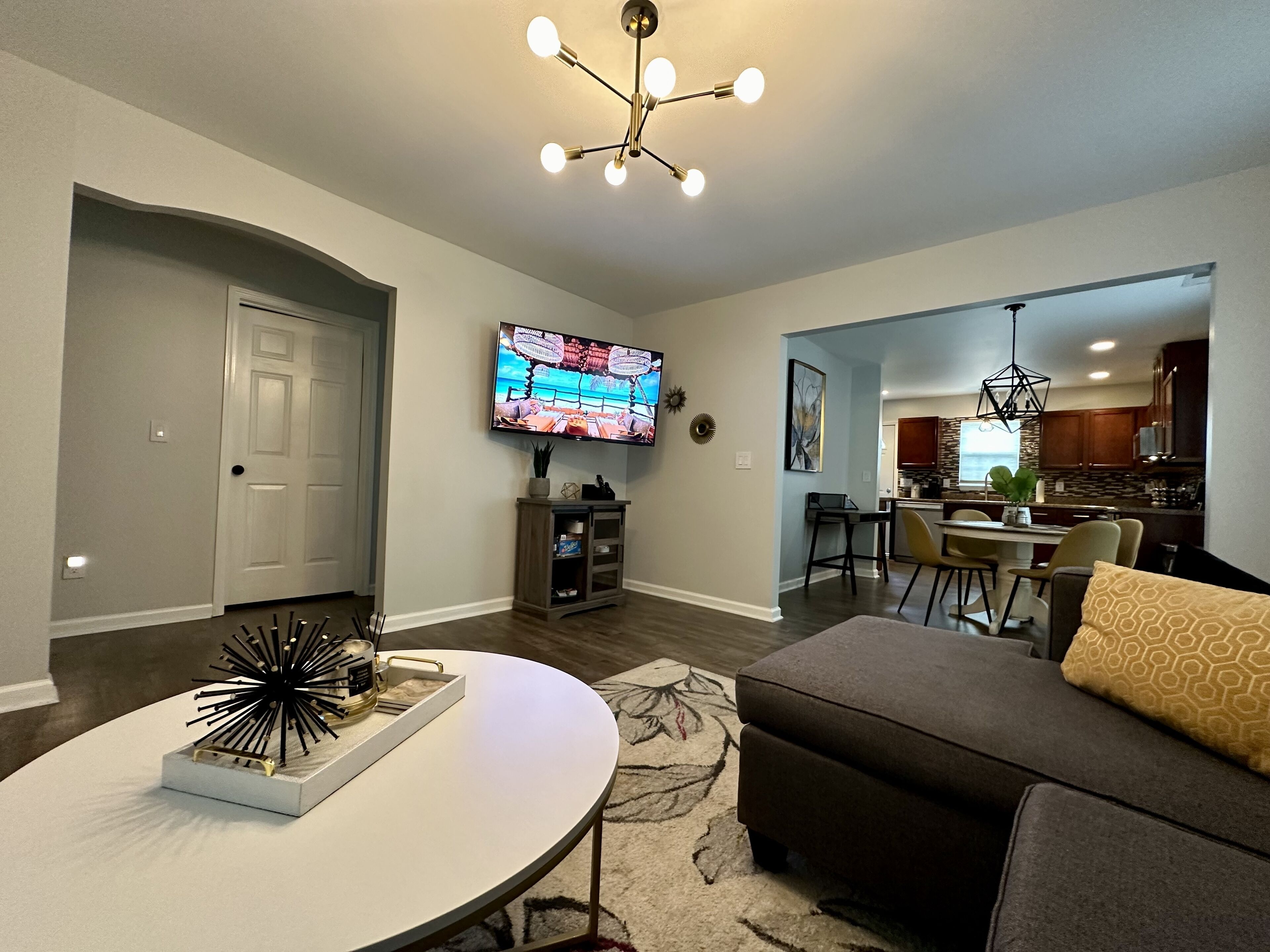 A modern living room with a flat-screen TV, a dining area, and a kitchen in the background.