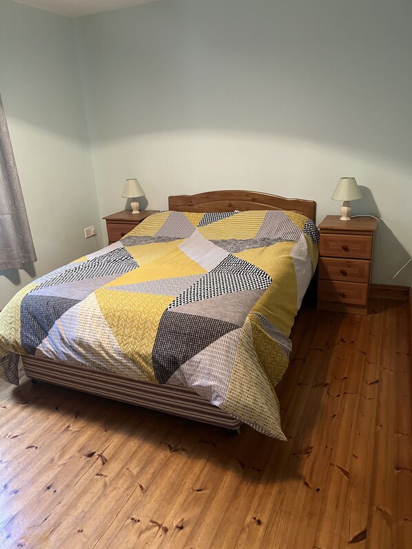 Een bed met een patchworkdeken, een houten hoofdbord, twee nachtkastjes met lampen en een houten vloer.