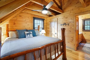 Een slaapkamer in een houten chalet, met schuine wanden, een bed met hoofdeinde, een plafondventilator en een raam met jaloezieën.