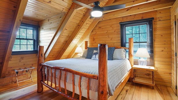 Quarto estilo loft com pé direito inclinado, cama com cabeceira de madeira, duas mesas de cabeceira com luminárias e janelas com persianas.