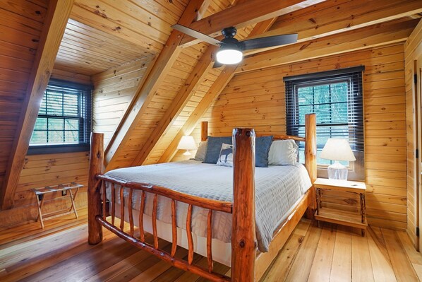 A wooden loft bedroom with a sloped ceiling, a bed with a wooden headboard, two bedside tables with lamps, and windows with blinds.