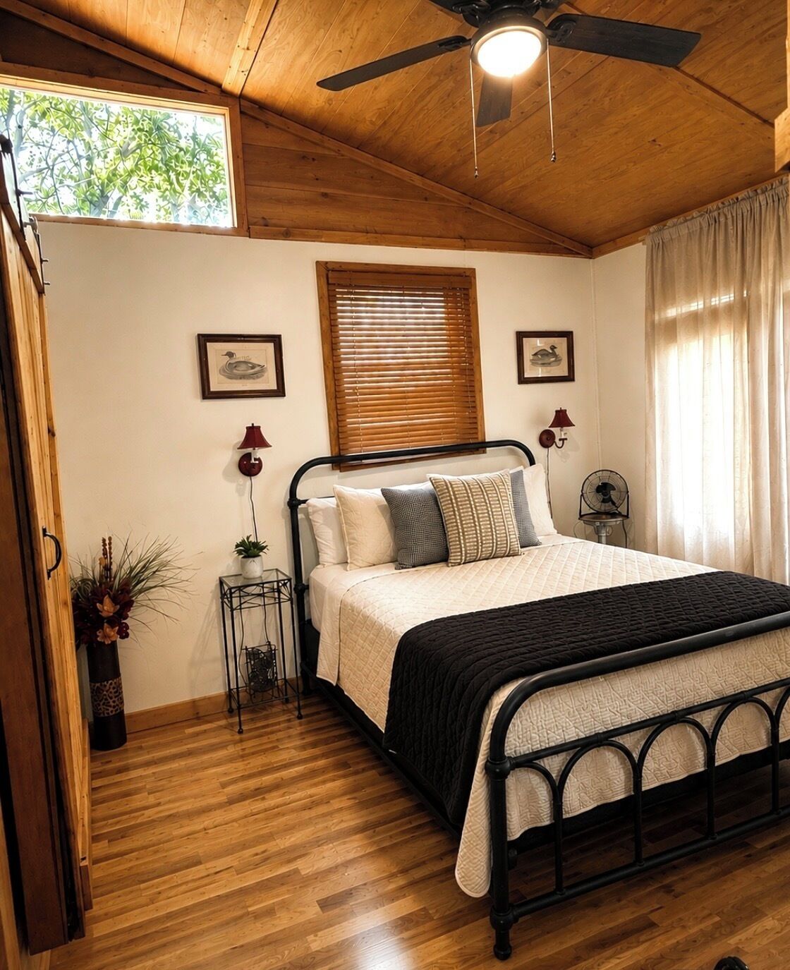 A bedroom with a bed, wooden floor, ceiling fan, window blinds, and a small side table with a lamp.