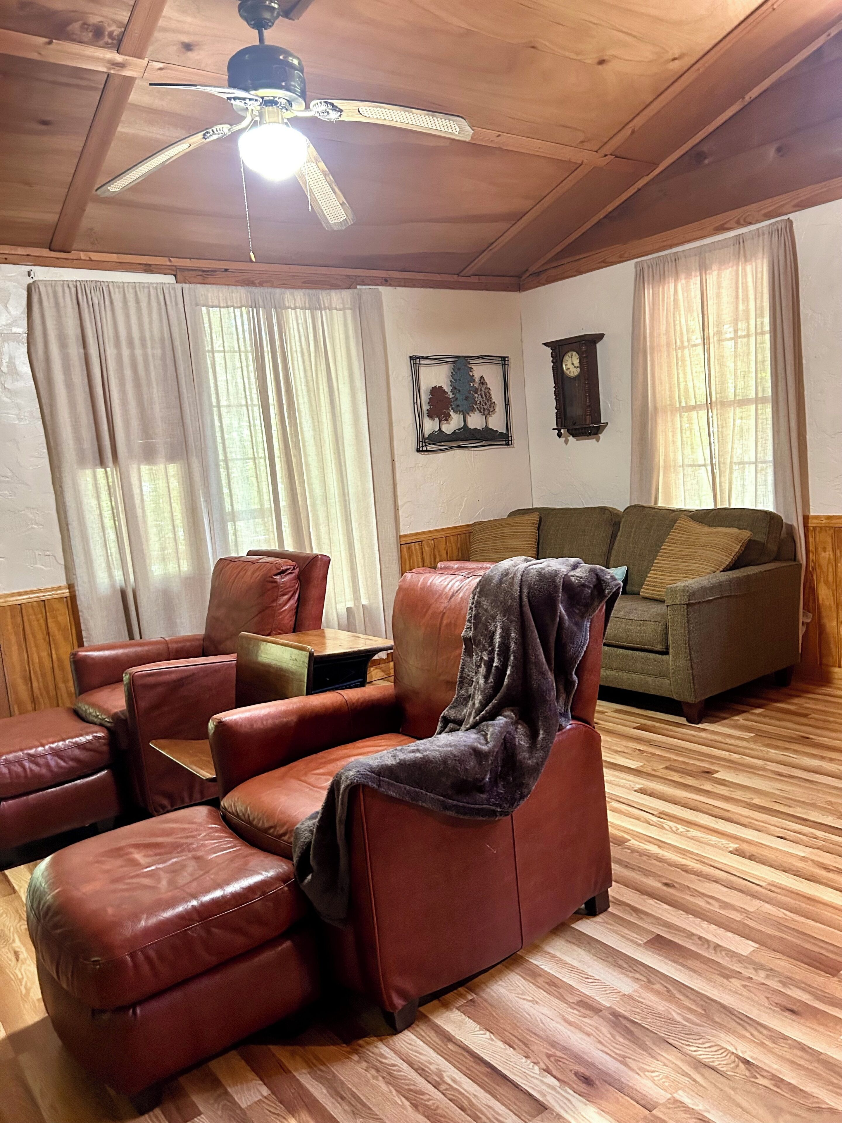 A room with a wooden ceiling, a ceiling fan, a brown leather sofa, a green armchair, and a wooden floor.