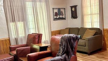 A room with a wooden ceiling, a ceiling fan, a brown leather sofa, a green armchair, and a wooden floor.