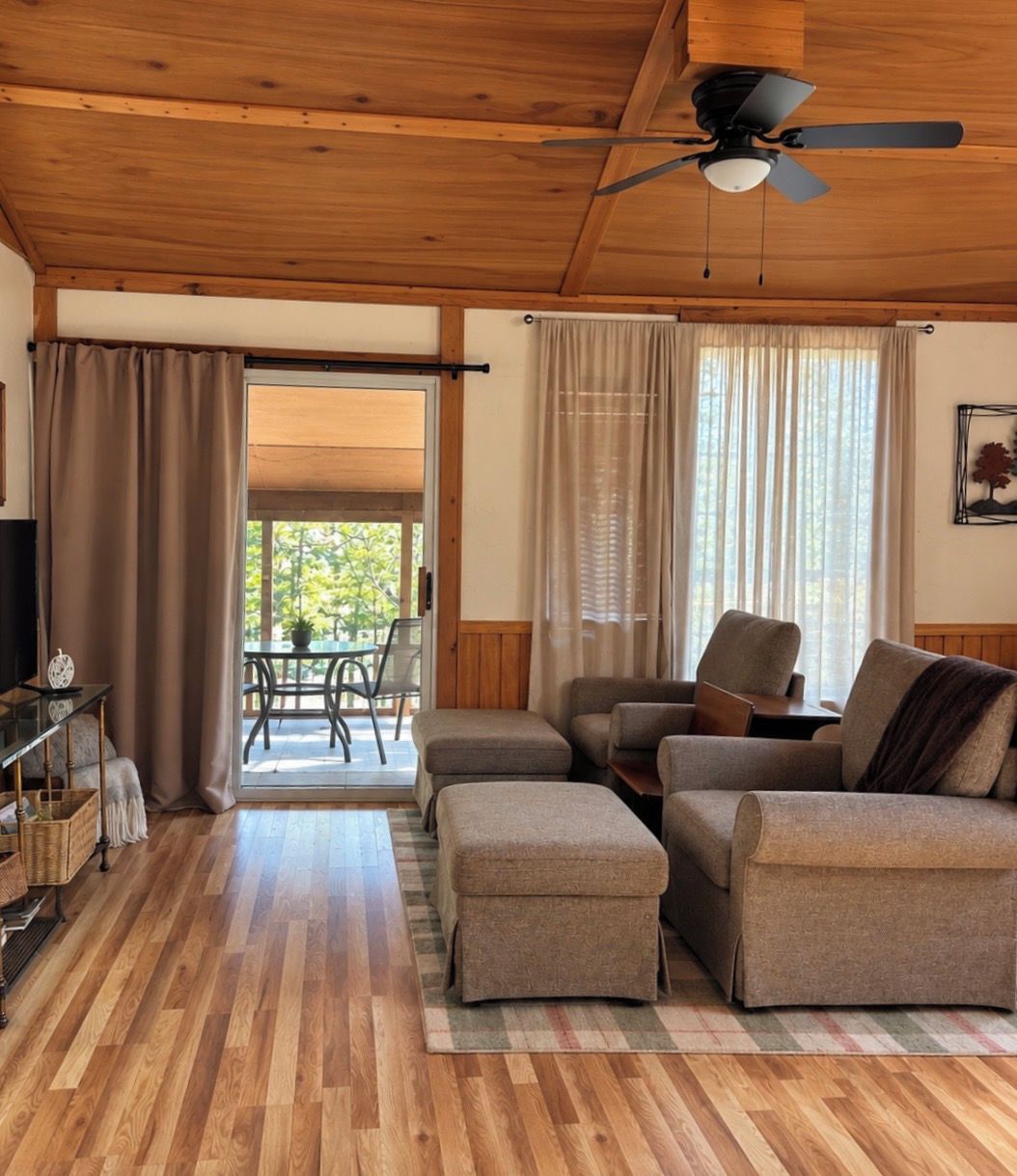 A room with a ceiling fan, wooden flooring, a sofa set, a television, and a view through a glass door to an outdoor area.