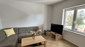 A living room with a grey sofa, a small wooden coffee table, a flat-screen TV on a stand, and a window with a view of greenery outside. - Villa Irmica - Impressive pool, spacious apartment (86 m²) - first floor (Donja Jagodnja)
