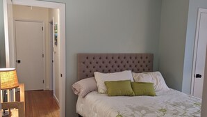 Een slaapkamer met een gestoffeerd hoofdeinde, een bed met bloemenbeddengoed, een houten vloer en een kamerkleed met een patroon.