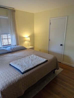 Un dormitorio con una cama, una mesita de noche con lámpara, una ventana con cortinas y una puerta.