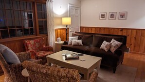Een gezellige woonkamer met een houten vloer, een bruine bank, rieten stoelen, een salontafel en een lamp.