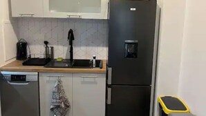 Une cuisine compacte avec des meubles blancs, un réfrigérateur noir, un lave-vaisselle et un pare-feu en carrelage.