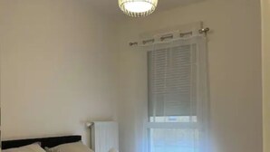 Une chambre à coucher comprenant un lit, des oreillers, une table de chevet, une lampe, une fenêtre avec des stores et un luminaire suspendu.