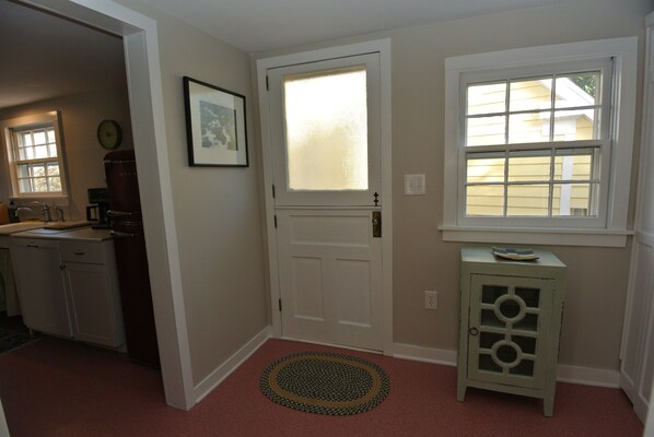 A hallway with a white door, a small cabinet with a glass door, a rug, and a window with blinds.