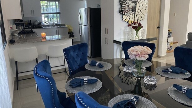 Un comedor moderno con una mesa de vidrio rodeada de sillas tapizadas en azul, un espejo decorativo y un jarrón con flores.