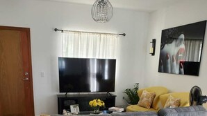 Una sala de estar con un sofá gris, un televisor de pantalla plana, una araña y un sofá amarillo.