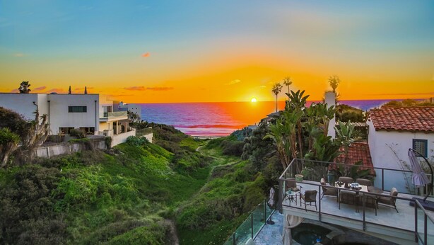 A coastal view with a sunset, a pathway leading to a building with outdoor seating, and a pool area.